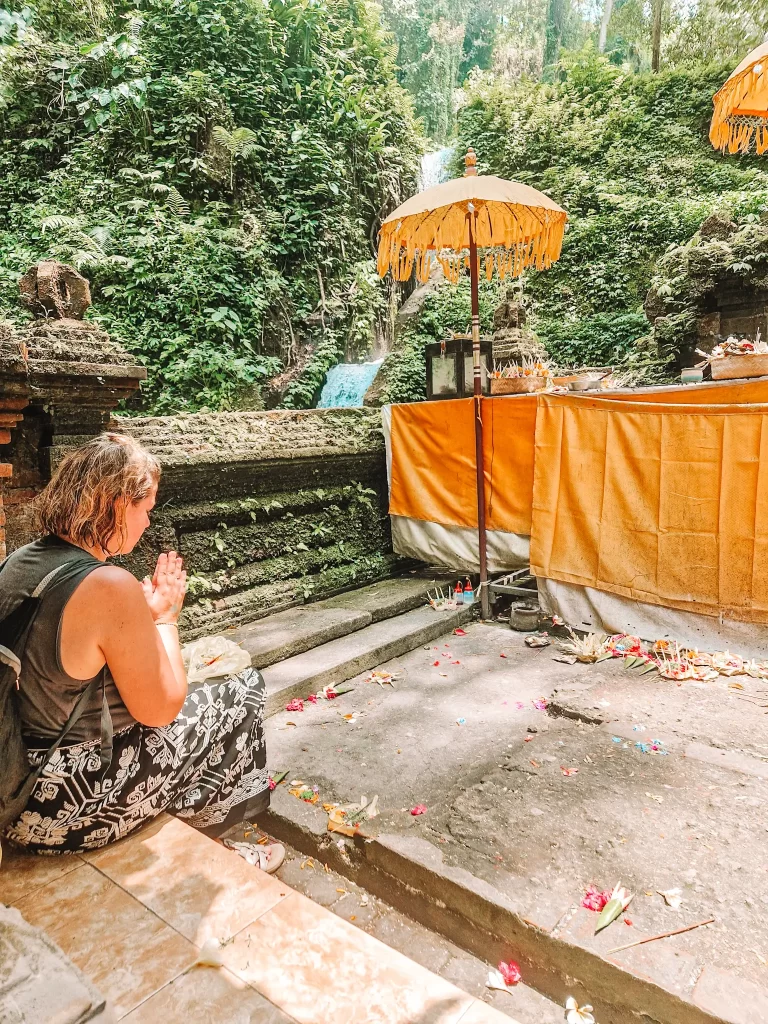 A woman dressed in a black sleeveless top and patterned sarong sits cross-legged at a Balinese water temple, pressing her hands together in prayer. She faces a sacred altar draped in yellow fabric, adorned with offerings and shaded by traditional umbrellas. Behind her, a lush green hillside with a cascading waterfall adds to the serene and spiritual atmosphere.