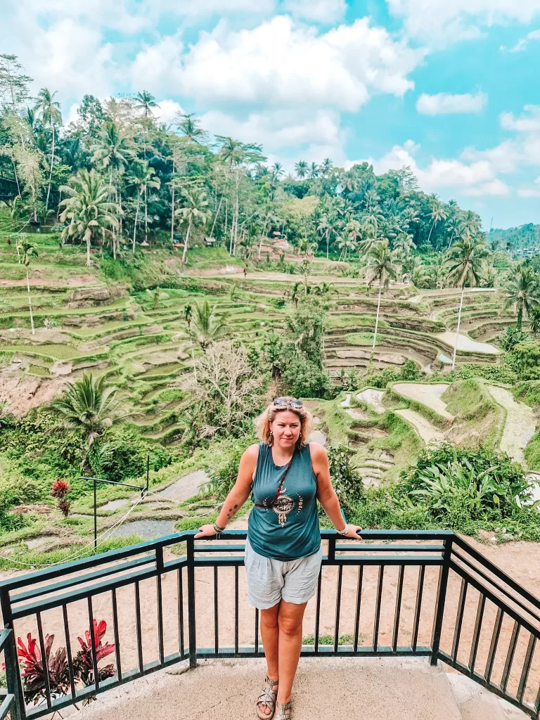 Em in a blue tank top and light shorts stands at a black metal railing, posing in front of Bali's iconic terraced rice fields. The lush green landscape stretches behind her, with tall palm trees and winding pathways weaving through the hills. The bright sky and tropical scenery create a picturesque backdrop.
