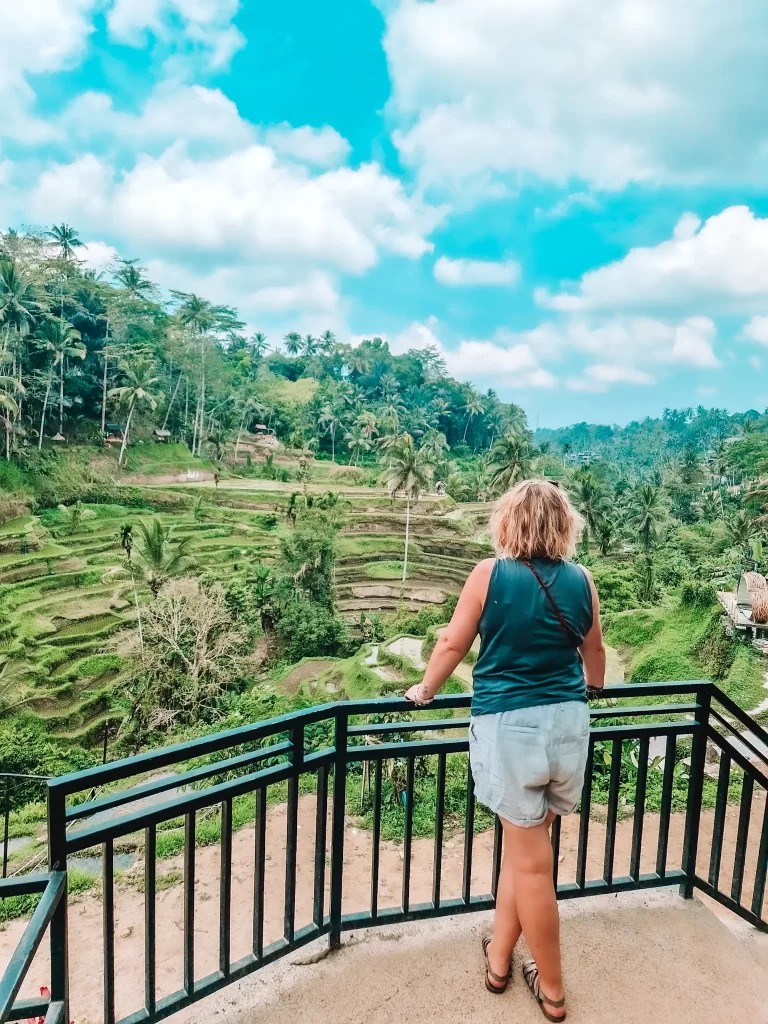 Em in a dark tank top and light shorts stands at a railing, gazing out at the lush, terraced rice fields of Bali. The landscape is filled with vibrant greenery, tall palm trees, and rolling hills under a bright blue sky with fluffy white clouds. The scene captures the peaceful beauty of rural Indonesia.