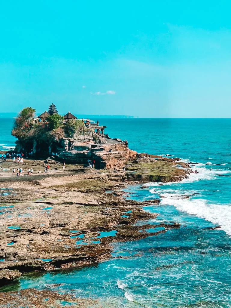 The iconic Tanah Lot Temple sits atop a rocky outcrop surrounded by turquoise ocean waves, with tourists exploring the shoreline under a bright blue sky.