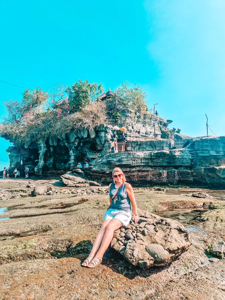 Em sits on a rock near the Tanah Lot temple in Bali, Indonesia, with the iconic sea temple towering behind her. She wears sunglasses, a sleeveless top, and shorts, looking relaxed as she enjoys the coastal scenery. The rocky shoreline and temple's rugged architecture contrast beautifully with the bright blue sky.