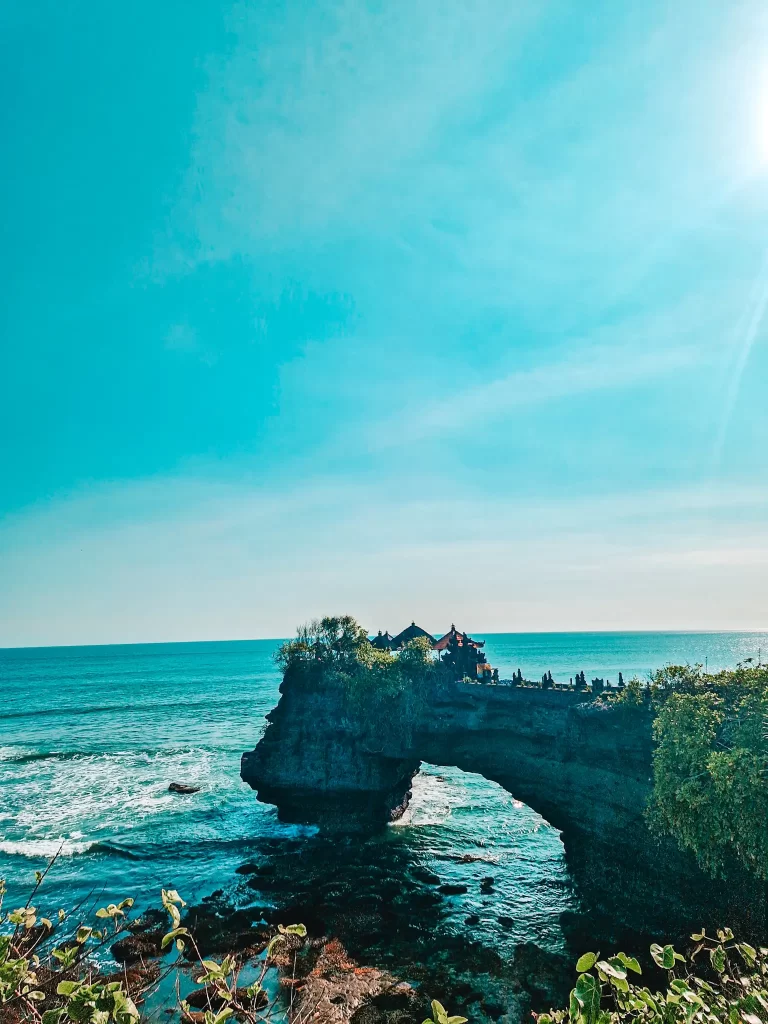 A stunning view of a Balinese sea temple perched atop a natural rock arch, surrounded by the deep blue ocean. The waves crash against the rocky shore as the temple stands silhouetted against the bright sky. Lush greenery frames the scene, adding to the serene and spiritual atmosphere.