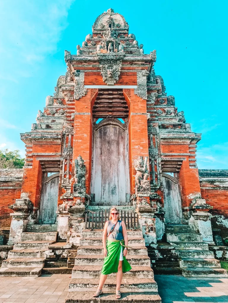 Em wearing sunglasses and a green sarong poses confidently on the stone steps of an ornate Balinese temple gate. The temple features intricate carvings and a weathered wooden door, framed by red brick and stone structures with guardian statues. The bright blue sky contrasts beautifully with the historic architecture, emphasizing the temple’s grandeur.