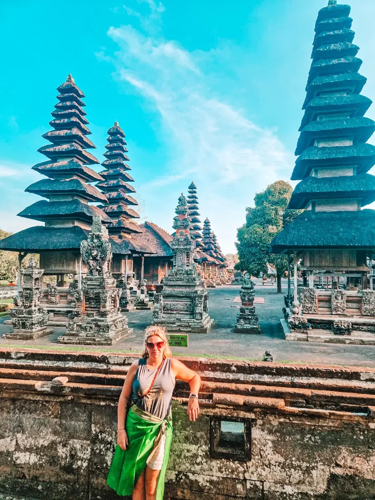 Em wearing a green sarong and sunglasses poses in front of the Taman Ayun Temple in Bali, Indonesia. The temple features towering multi-tiered pagodas, intricate stone carvings, and a peaceful courtyard. The warm sunlight highlights the ancient architecture, creating a serene and cultural atmosphere.