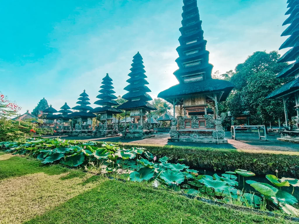 A stunning view of Taman Ayun Temple in Bali, Indonesia, showcasing its multi-tiered pagoda-style shrines. The temple's intricate stone carvings and traditional Balinese architecture stand against a bright blue sky. A lush lotus pond and well-manicured greenery add to the serene and sacred atmosphere.