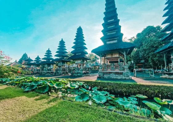 A stunning view of Taman Ayun Temple in Bali, Indonesia, showcasing its multi-tiered pagoda-style shrines. The temple's intricate stone carvings and traditional Balinese architecture stand against a bright blue sky. A lush lotus pond and well-manicured greenery add to the serene and sacred atmosphere.