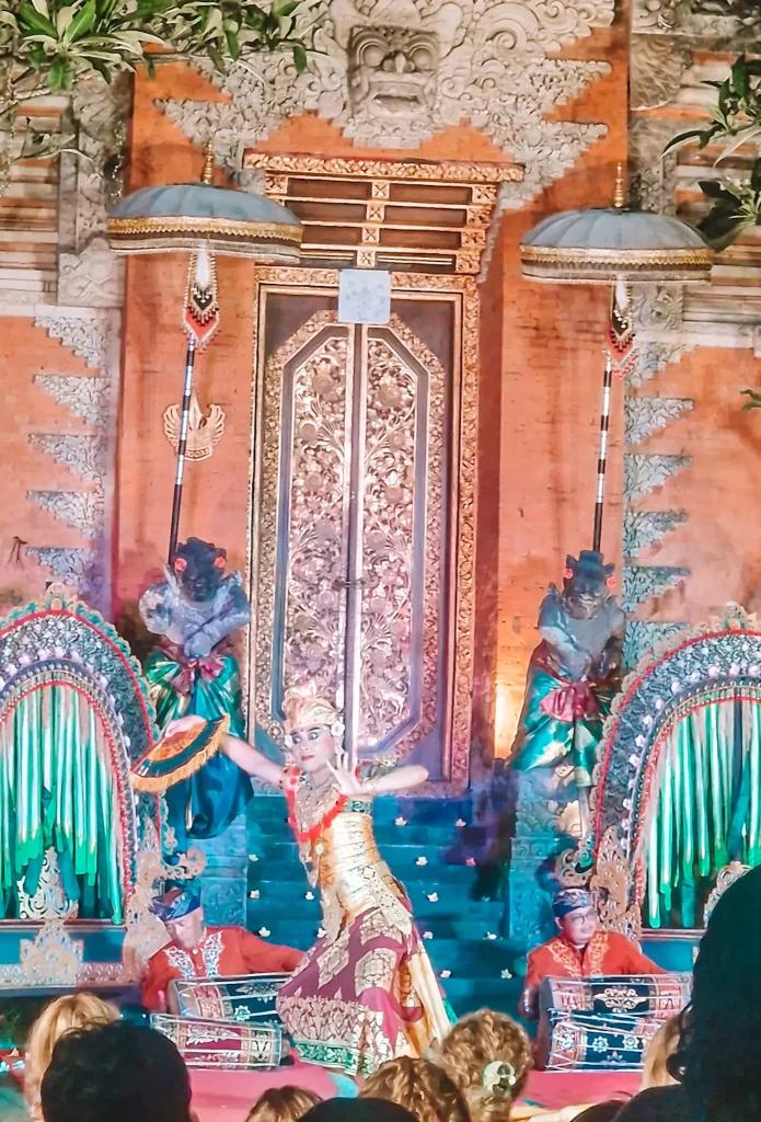 A Balinese dancer in an ornate gold and red costume gracefully performs a traditional dance in front of a richly decorated temple gate. Her poised movements and expressive gestures are illuminated by warm lighting, while musicians in red attire play gamelan instruments in the background. The intricate carvings and ceremonial umbrellas add to the cultural ambiance of the performance.