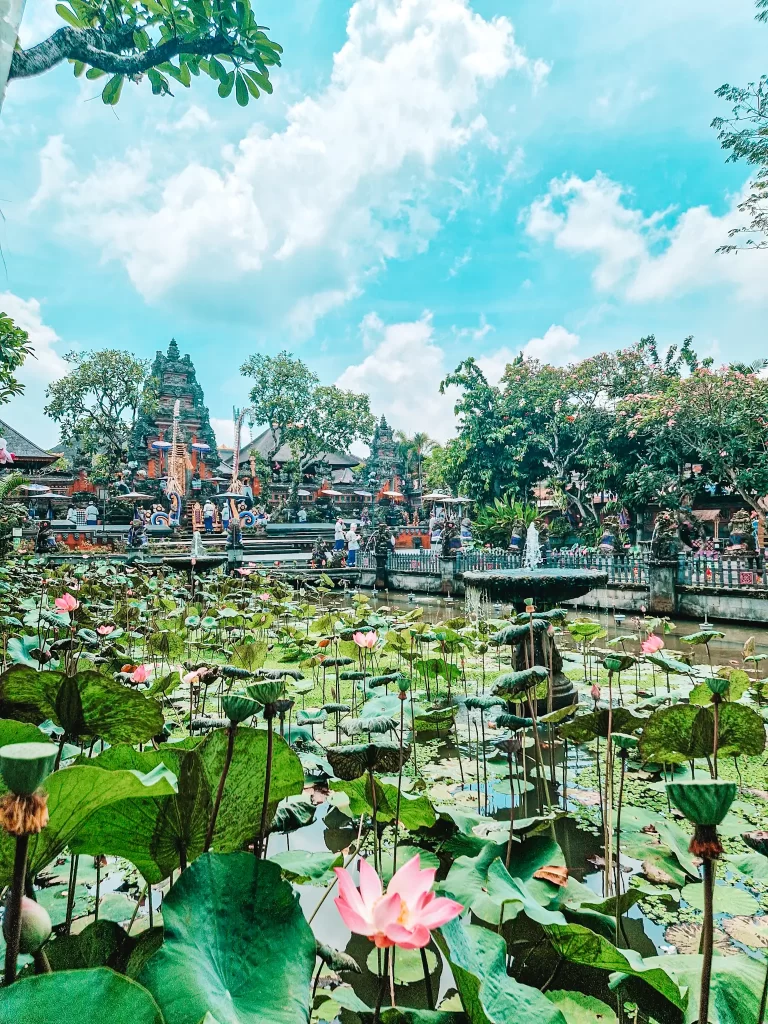 A serene lotus pond filled with vibrant pink blossoms stretches across the foreground, leading to an intricately designed Balinese temple in the background. The temple’s traditional architecture, with detailed carvings and statues, is surrounded by lush greenery under a bright blue sky with fluffy clouds. Visitors can be seen exploring the sacred site, adding to the peaceful yet lively ambiance.