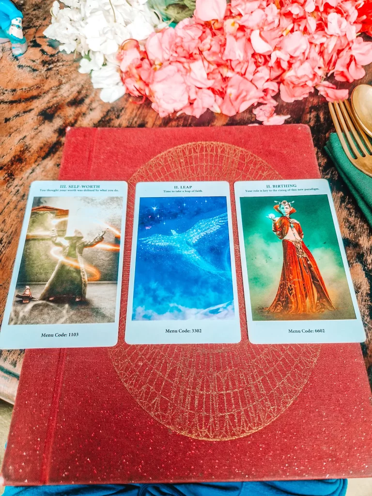 Three oracle or tarot cards are laid out on a textured red journal with a gold geometric design, placed on a wooden table. The cards read: "III. Self-Worth – You thought your worth was defined by what you do," "II. Leap – Time to take a leap of faith," and "II. Birthing – Your role is key to the rising of this new paradigm." Behind the cards, pink and white flower petals are scattered, adding a mystical and decorative touch.