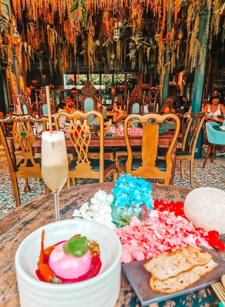 A beautifully decorated café with intricate wooden chairs and hanging vines, featuring a table with a tropical cocktail, a vibrant floral arrangement, and a plated dish with pink mousse, roasted vegetables, and crispy bread.