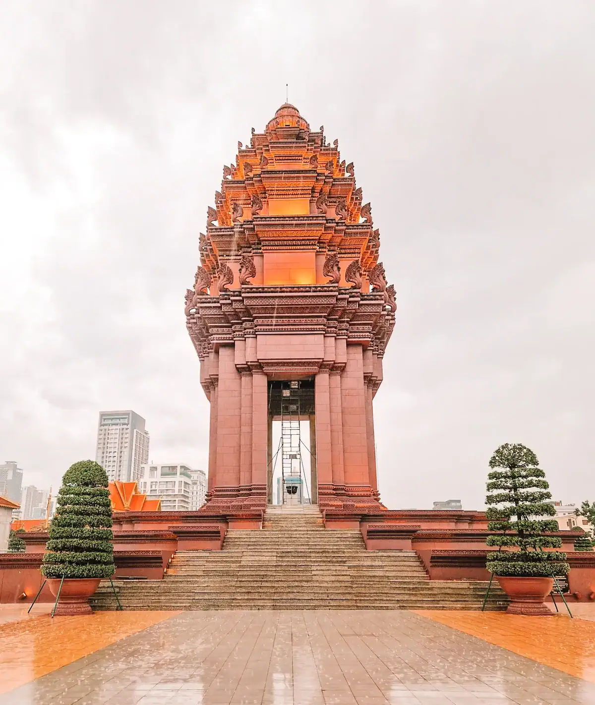 A towering, intricately carved monument made of reddish-brown stone, resembling traditional Khmer temple architecture. The monument stands on a raised platform with stairs leading up to an open archway, revealing a bell inside. Two neatly trimmed, spiral-shaped trees flank the monument, with a modern city skyline visible in the background under an overcast sky.