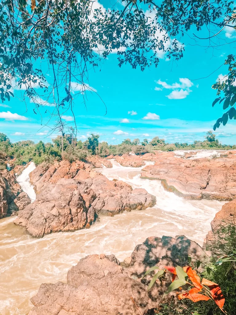 A river crashes over jagged, reddish-brown rocks, creating a dramatic waterfall scene. Green foliage frames the top and bottom of the image, while the sky is a vivid blue with scattered clouds.