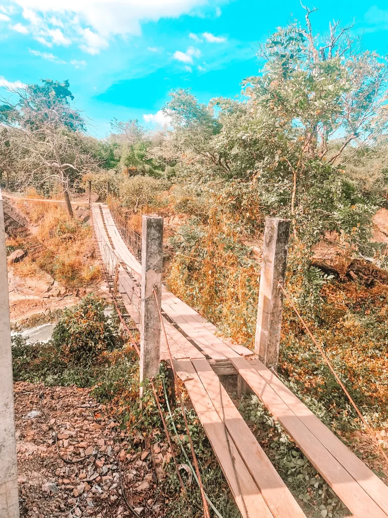 A rustic wooden suspension bridge with planks and rope railings stretches across a small river, surrounded by lush greenery and dry, golden foliage. The sky is bright blue with scattered clouds, adding a vibrant contrast to the earthy tones of the landscape. The bridge appears weathered and slightly uneven, blending naturally into the rural setting.