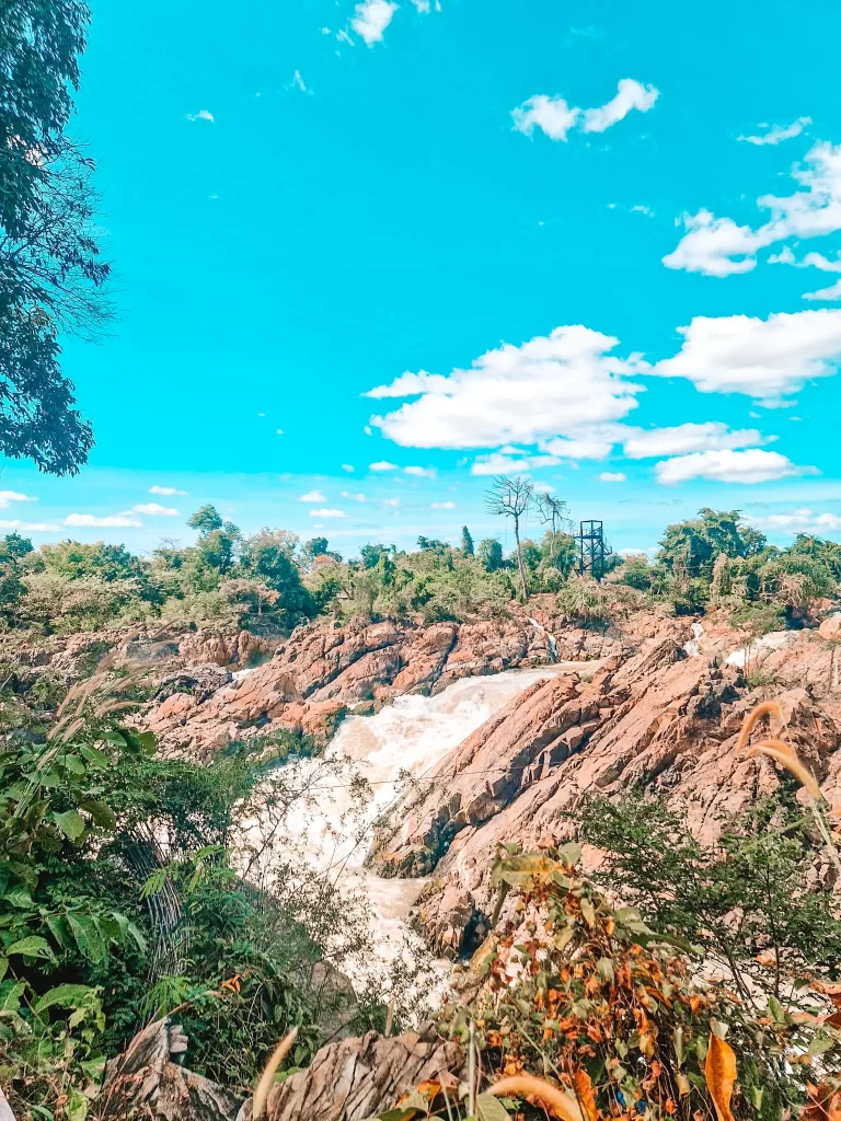 A rushing waterfall cascades down rugged, reddish-brown rocks, surrounded by lush green foliage. The bright blue sky is dotted with fluffy white clouds, adding to the vibrant and natural beauty of the scene.