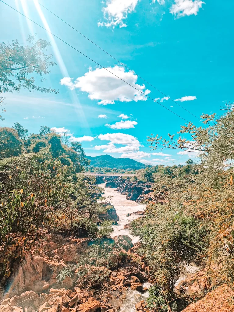 A powerful river cuts through a rocky canyon, surrounded by green vegetation. Sunlight beams down from the sky, casting a golden glow over the scene, with distant mountains visible on the horizon.