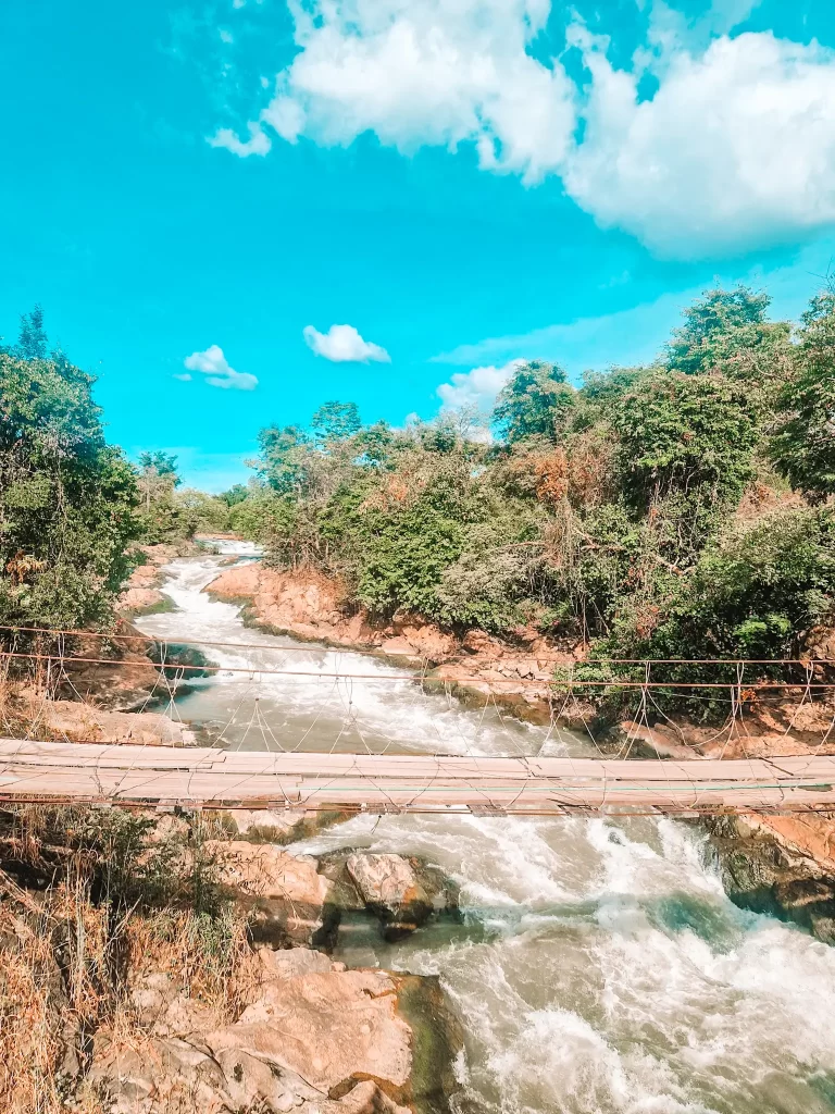 A wooden suspension bridge stretches over a fast-flowing river with white rapids. Dense green trees line the riverbanks, and the sky above is a brilliant blue with scattered clouds.