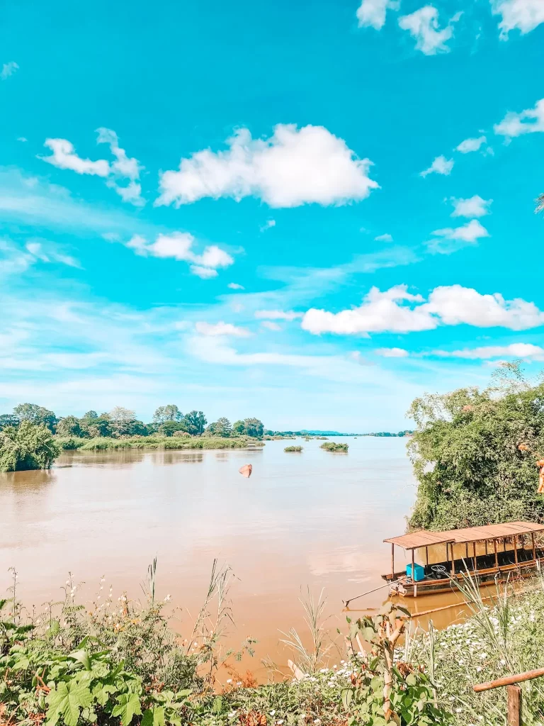 A serene river under a vibrant blue sky with fluffy white clouds. A wooden boat with a thatched roof is docked by the riverbank, surrounded by lush greenery.