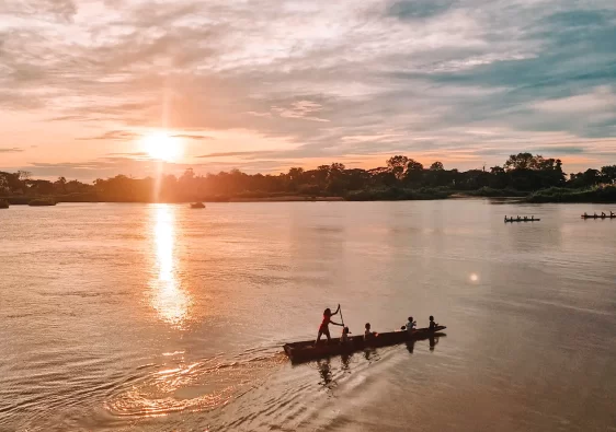 Children paddle a long wooden boat carrying several passengers across a river at sunrise. The golden light reflects on the water, creating a shimmering path. In the background, another boat with rowers moves in the same direction.