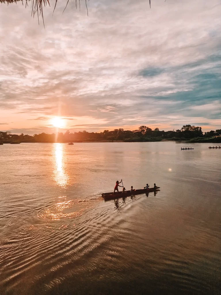 Children paddle a long wooden boat carrying several passengers across a river at sunrise. The golden light reflects on the water, creating a shimmering path. In the background, another boat with rowers moves in the same direction.