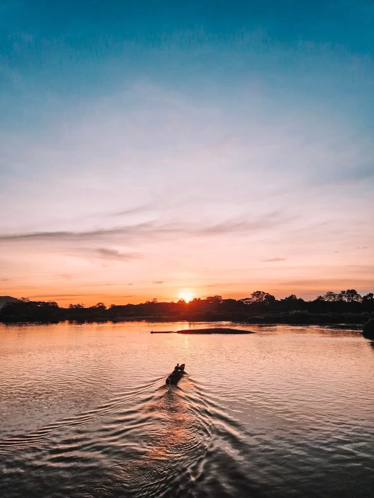 A wooden boat moves through a calm river at sunset, leaving gentle ripples behind. The sky is a blend of warm orange and soft blue hues, reflecting beautifully on the water. Silhouettes of trees and distant hills line the horizon.