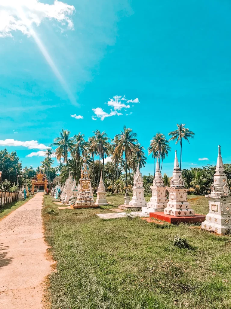 A row of ornate, white and gold stupas stands in a lush green field under a vivid blue sky. Palm trees line the background, and a golden temple entrance is visible in the distance. Sunlight streams through the clouds, casting a peaceful glow.