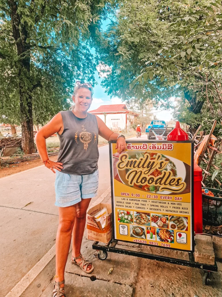 A woman in casual summer clothes stands next to a sign for "Emily’s Noodles" street food stall, smiling at the camera. The sign advertises various dishes, including noodle soup, fried rice, and spring rolls.