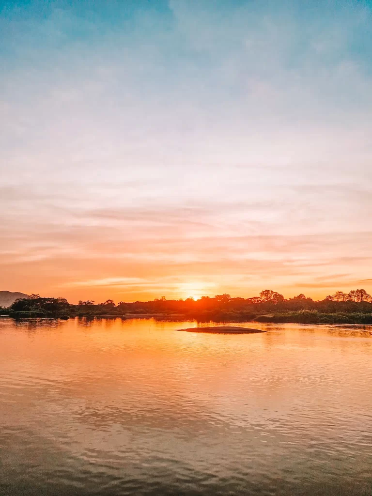 A tranquil river reflects the warm hues of the setting sun, with a small island visible in the distance. The sky blends soft orange and blue tones, creating a serene sunset scene.