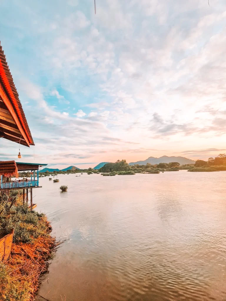 A riverside scene at sunset, with a stilted wooden structure extending over the water. The golden light illuminates the landscape, casting a glow on the distant mountains and small islands scattered across the river.