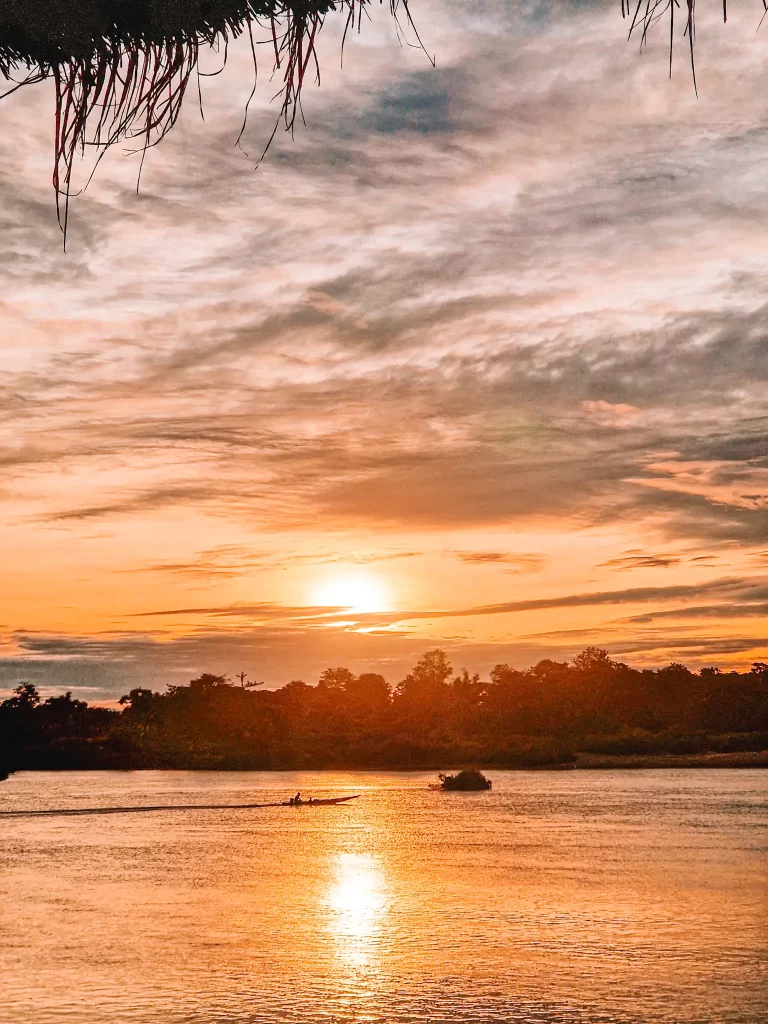 A golden sunset reflects on the calm waters of a river, with a lone boat moving across the water. Silhouettes of trees line the horizon, and strands of thatched roofing frame the top of the image.