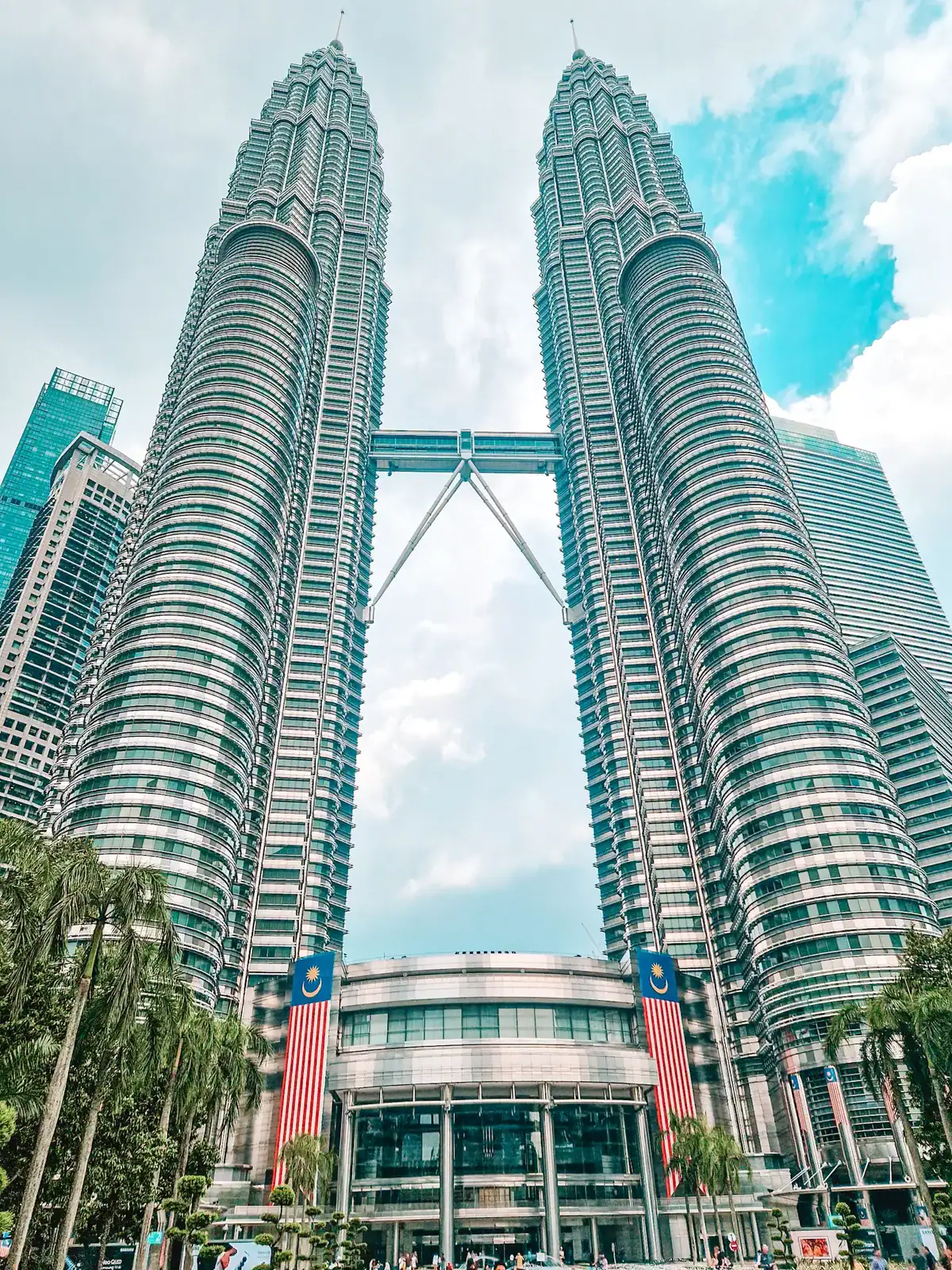 A low-angle view of the Petronas Twin Towers in Kuala Lumpur, Malaysia, with their sleek silver facade and skybridge connecting the two structures. The foreground features palm trees and two large Malaysian flags hanging from the entrance.