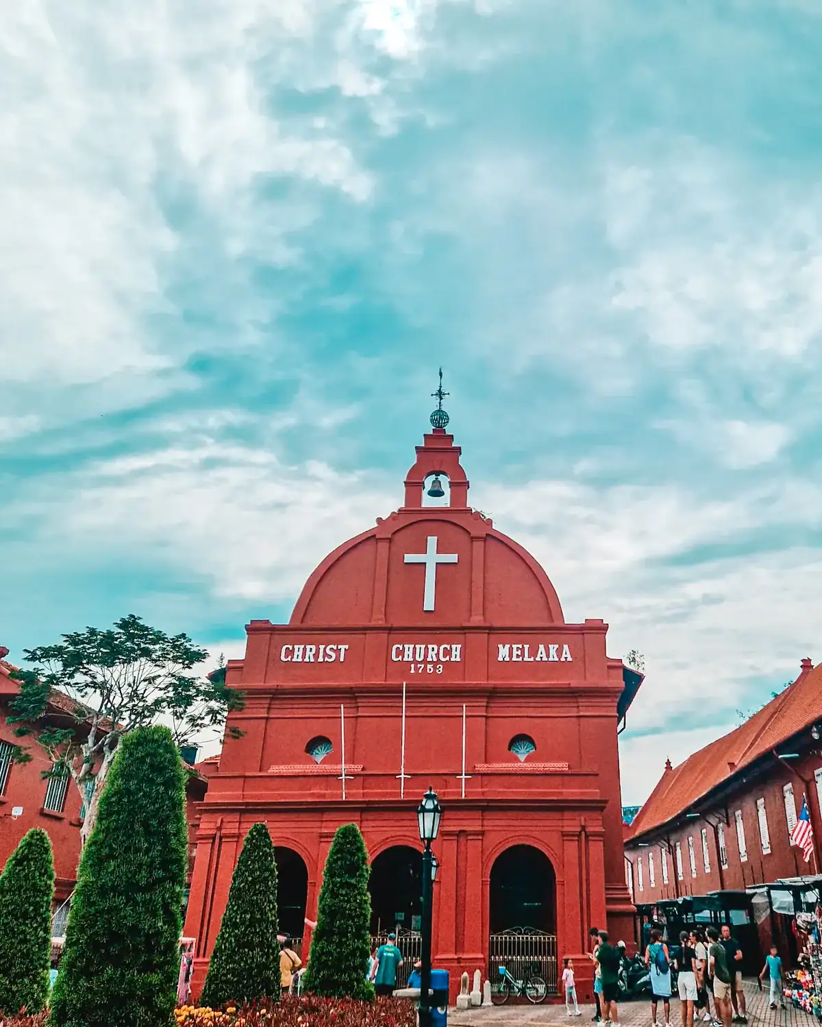Christ Church Melaka, a historic red-brick church built in 1753, stands against a blue sky. The church’s white cross and lettering contrast with its deep red facade, while visitors walk around the square, surrounded by neatly trimmed greenery.