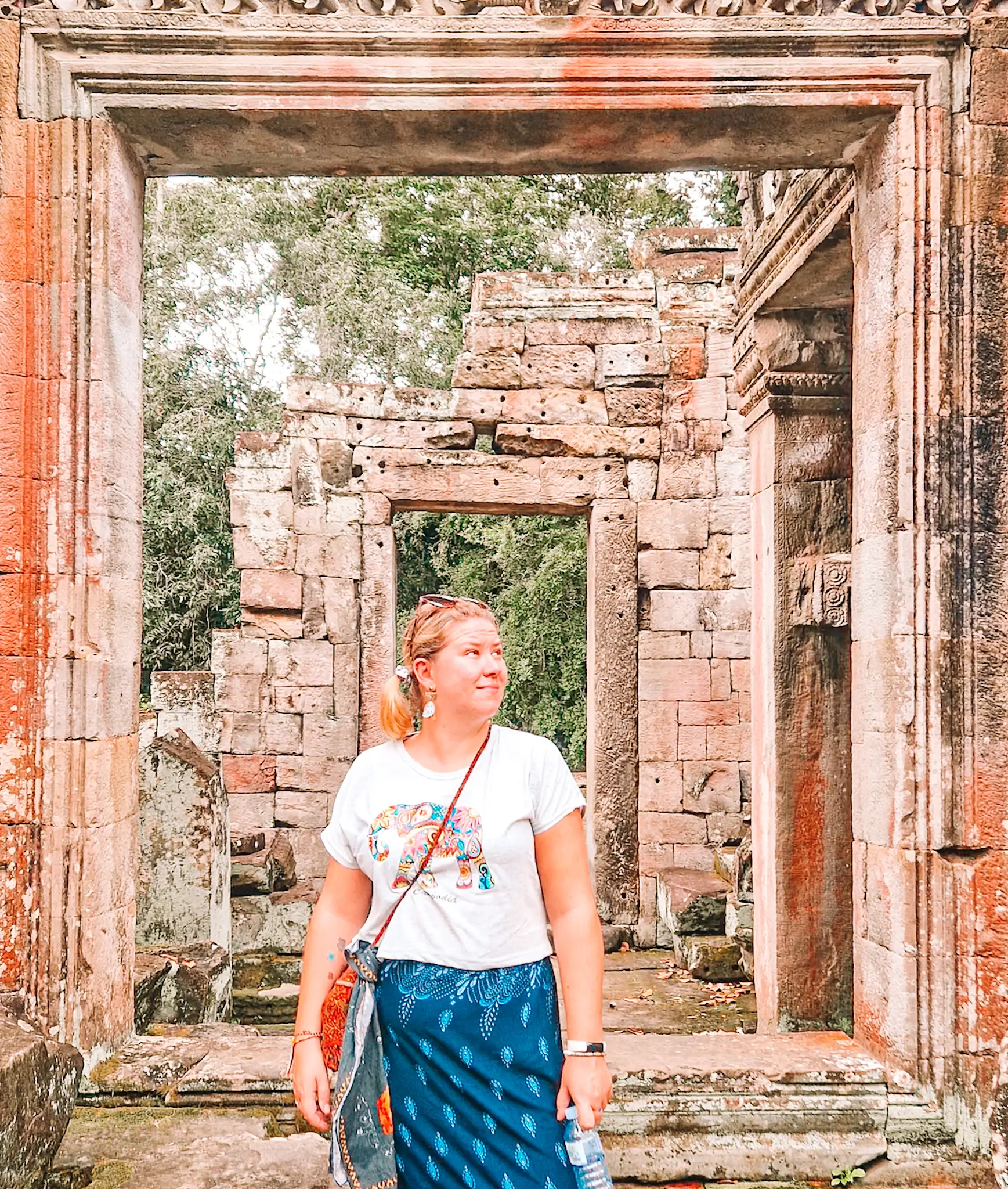 A woman with blonde hair tied back in a ponytail stands in front of an ancient stone temple ruin, framed by a large rectangular doorway. She wears a white t-shirt with a colorful elephant design, a blue patterned skirt, and carries a crossbody bag. She gazes off to the side with a relaxed expression, while the weathered stone walls and lush greenery in the background add to the historic ambiance.