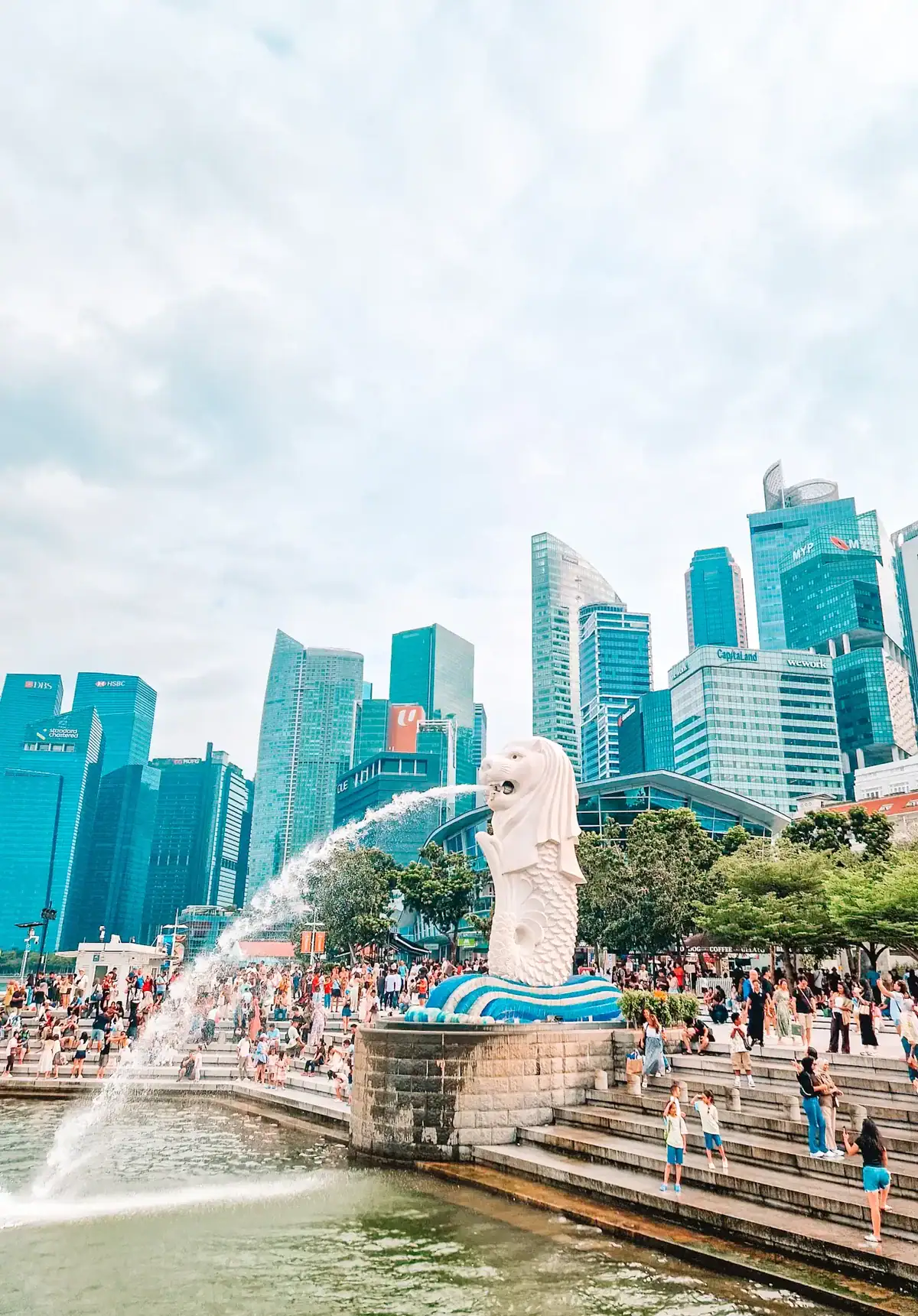 The iconic Merlion statue in Singapore, a white half-lion, half-fish figure, spouts water into the bay. A bustling crowd gathers around the waterfront, with modern skyscrapers forming the city skyline in the background.