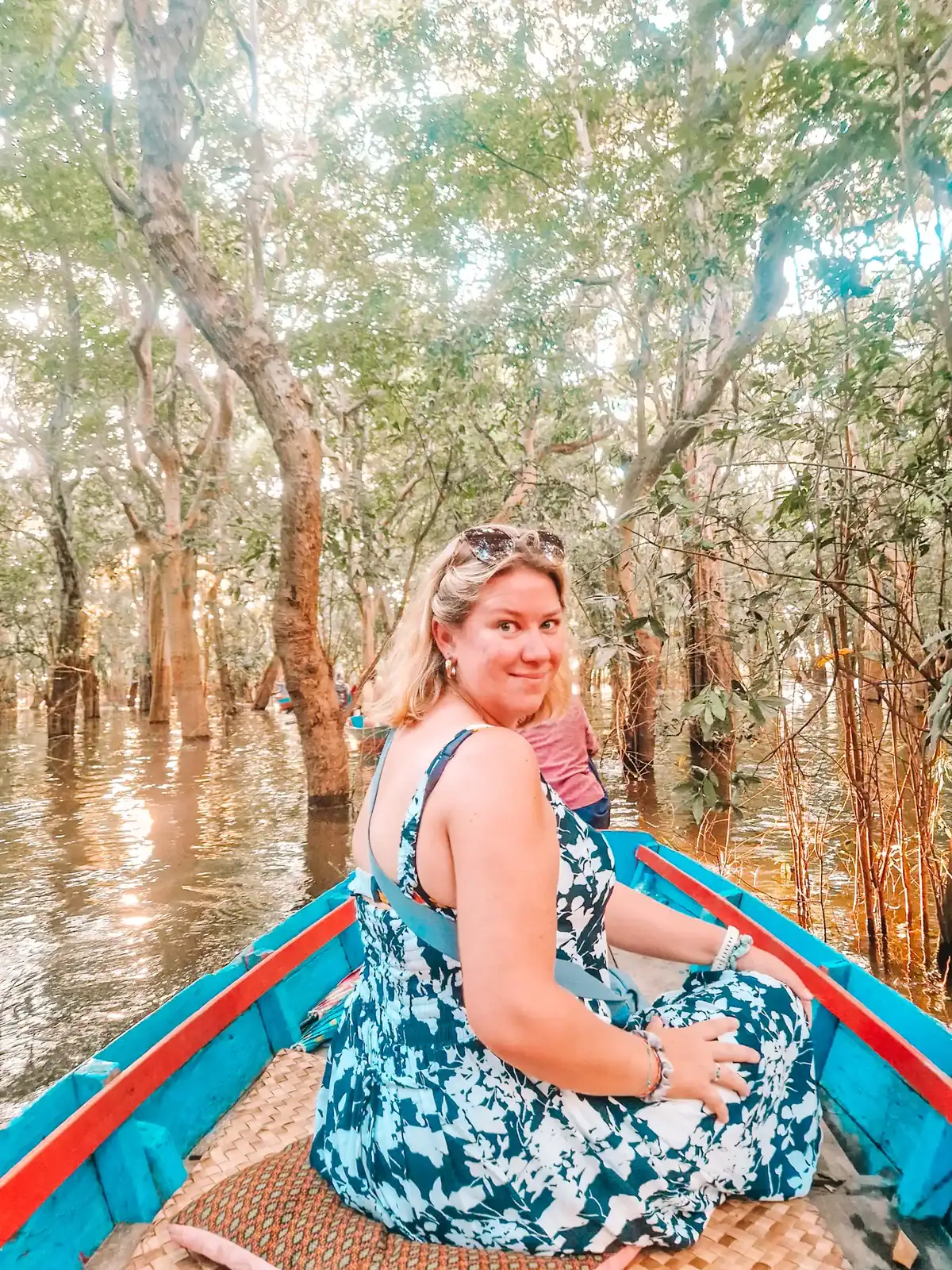 A woman in a blue and white floral dress sits on a blue wooden boat, smiling at the camera as she navigates through a flooded forest. Sunlight filters through the dense green canopy, reflecting on the water. Trees with partially submerged trunks surround the boat.