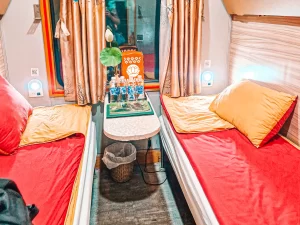 A cozy train cabin with two parallel beds covered in red and yellow bedding, a small table in between with bottled water and a lotus flower, and soft lighting illuminating the space.