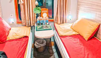 A cozy train cabin with two parallel beds covered in red and yellow bedding, a small table in between with bottled water and a lotus flower, and soft lighting illuminating the space.