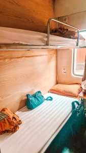 Interior of a compact train sleeper cabin with two bunk beds, orange pillows, and bags placed on the lower bunk. There's a small window bringing in natural light and showing a glimpse of a train platform outside.