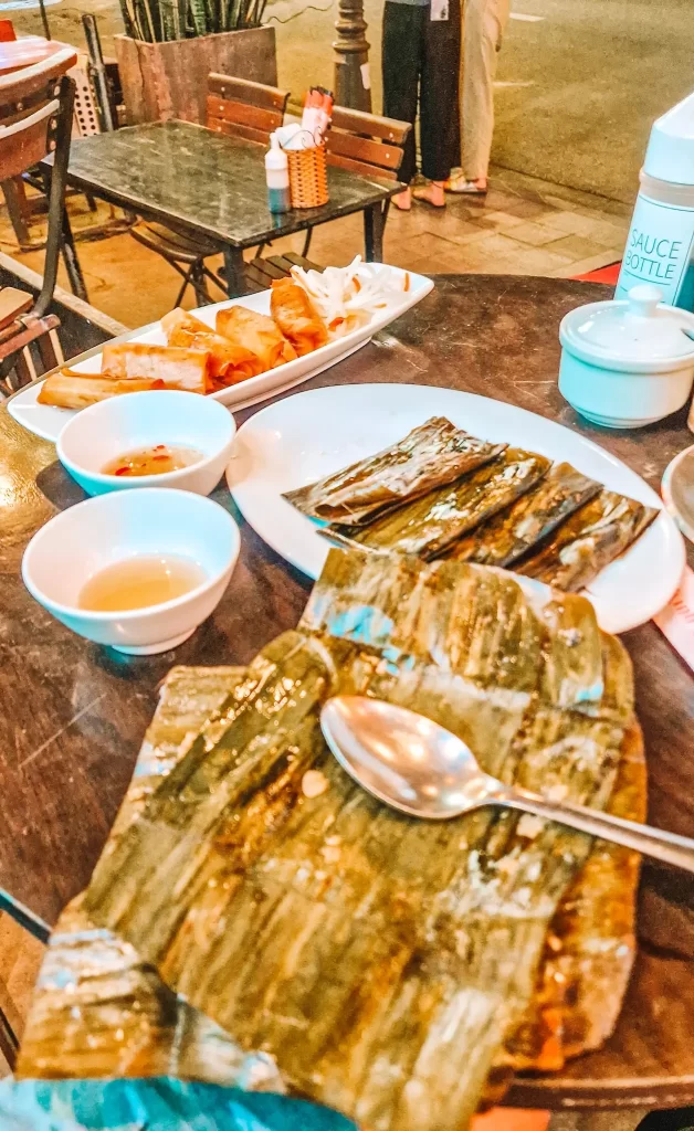 Plate of vegan food, with unwrapped banana leaves at the front and wrapped banana leaf parcels behind. There are two small bowls of sauce, and a plate of fried spring rolls. All are on a wooden table, with another wooden table visible in the background. 