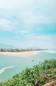 Scenic coastal landscape featuring turquoise water, a sandy beach stretching along the shore, and a small boat floating near the coastline. In the background, a village and misty mountains rise under a soft blue sky.
