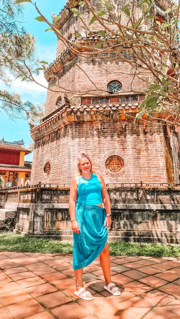 Woman in a blue dress smiling in front of the brick facade of Thien Mu Pagoda, soaking in the history and architecture during 2 days in Hue.