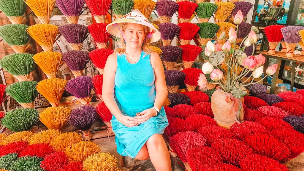 Woman wearing a conical hat and sitting among colorful bundles of incense sticks at a traditional market, a vibrant cultural experience to enjoy during 2 days in Hue.