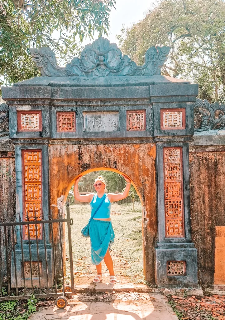 A woman in a blue outfit poses under a weathered stone archway decorated with intricate patterns, capturing the charm of hidden historical corners during 2 days in Hue.