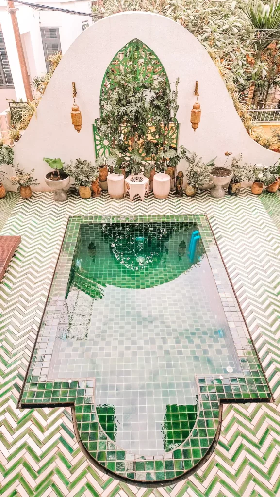 Small, green-tiled plunge pool with a decorative geometric shape, surrounded by patterned green-and-white flooring and tropical plants along a curved wall with ornate green latticework.