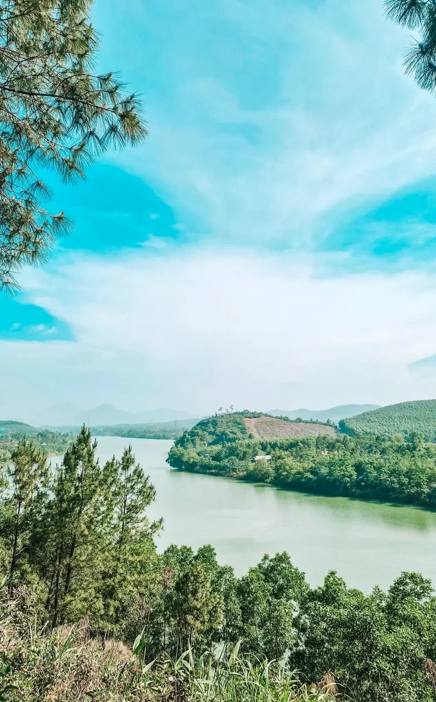 Panoramic view of the Perfume River winding through lush green hills and pine trees under a hazy sky, a serene sight often enjoyed during 2 days in Hue.
