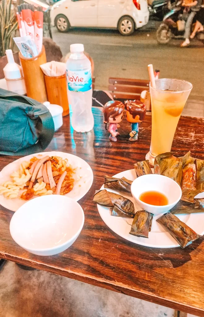 Wooden table with multiple plates of food, including banana leaf-wrapped parcels surrounding a bowl of sauce, and a plate with pasta and vegan ham. There is an orange drink behind, with two small dolls, a bottle of water and a handbag on the table.