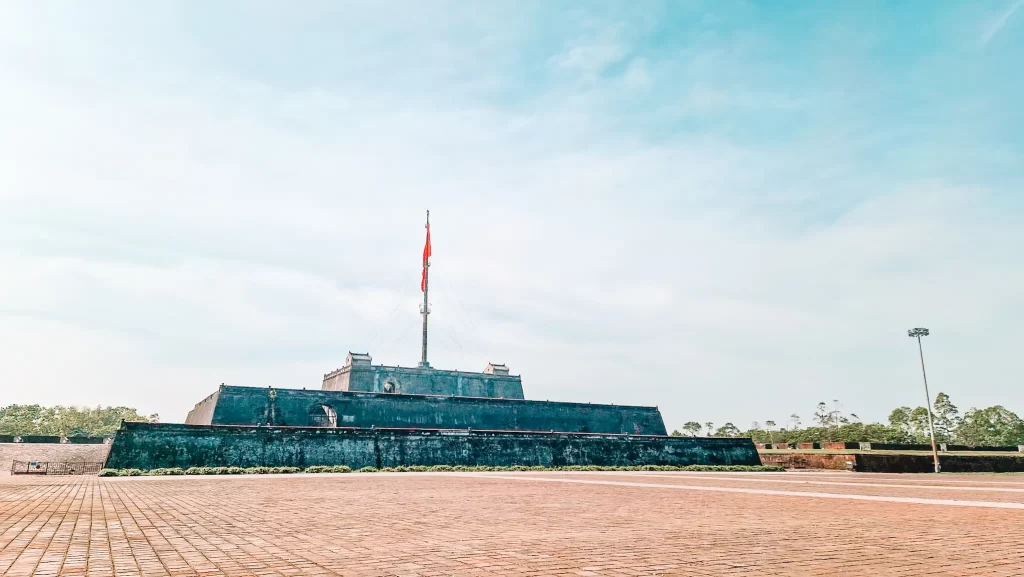 Wide shot of the giant Flag Tower at the Imperial Citadel with a bright red flag waving under a pale blue sky, a symbolic landmark to visit during 2 days in Hue.