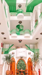 Open-air hotel atrium with white walls and vibrant green railings, showcasing multiple floors connected by a green spiral staircase. The sign above the entrance reads “Pisces Hotel,” flanked by potted plants and wooden archways.
