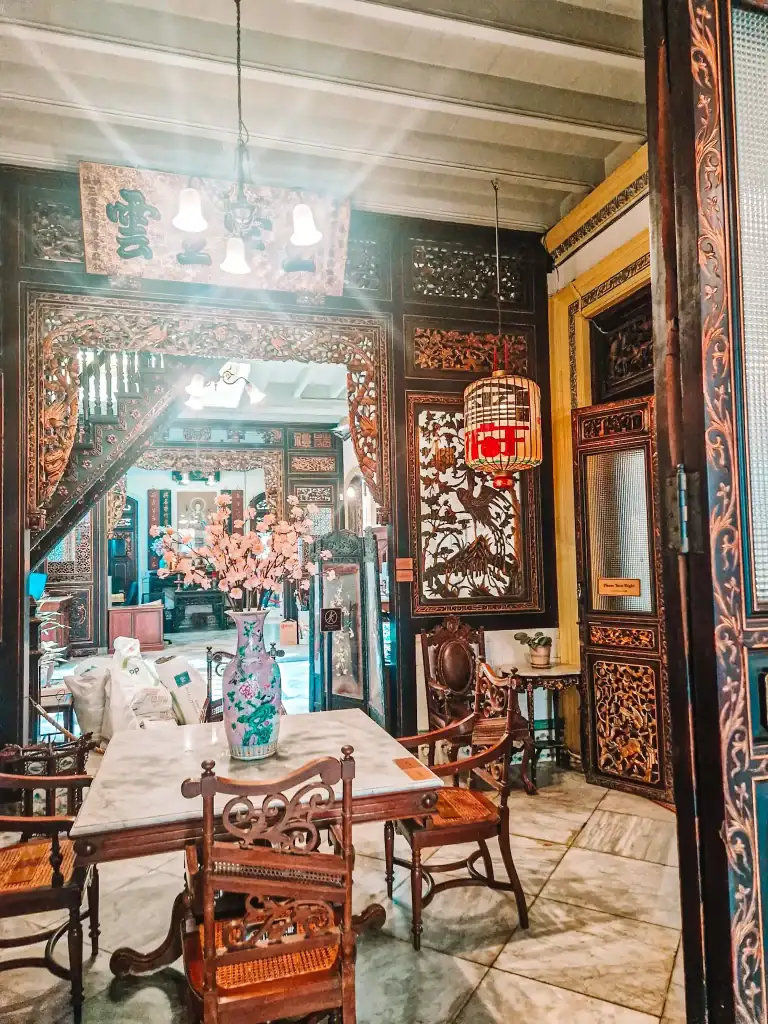 Ornate interior of a heritage Chinese mansion featuring intricate wood carvings, a marble-top dining table with antique chairs, and a tall porcelain vase with pink blossoms as the centerpiece. Traditional hanging lanterns and detailed paneling add to the room’s rich, historic elegance.