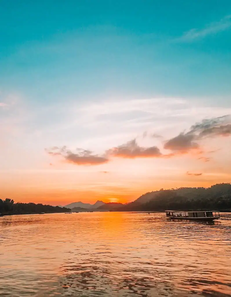 A golden sunset reflects over the Mekong River as a traditional longboat drifts by, capturing one of the most serene and iconic moments among the best things to do in Luang Prabang.