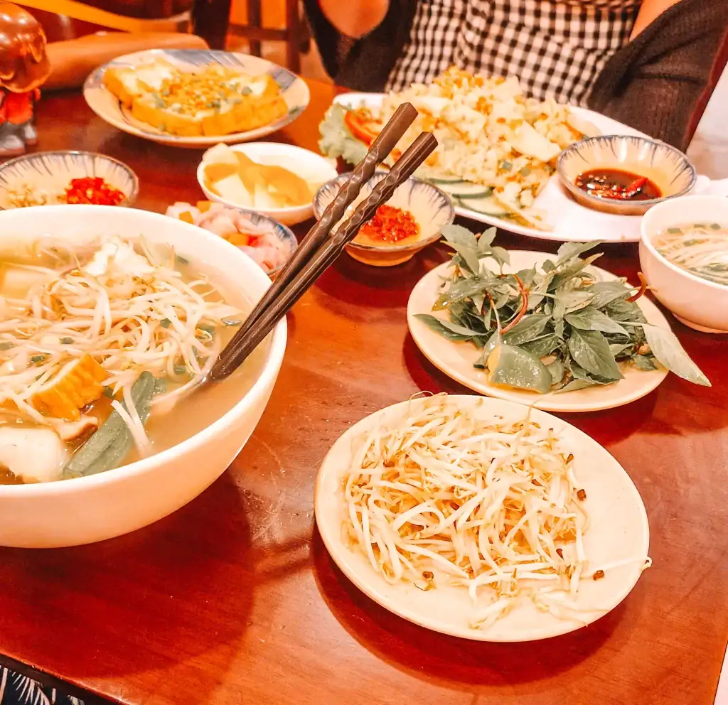 A range of plates on a table, including a big bowl of vegetable pho, beansprouts, various plates of sauces and herbs, and a plate of fried rice, all on a wooden table.